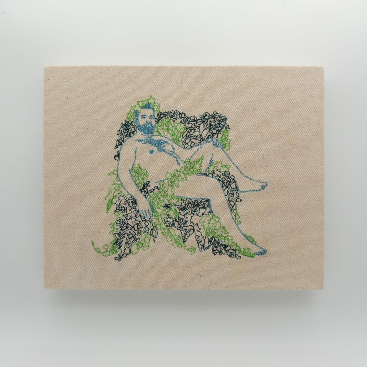 Marin: embroidered figure with a sparkly light blue beard, reclining in green leaves, on natural canvas